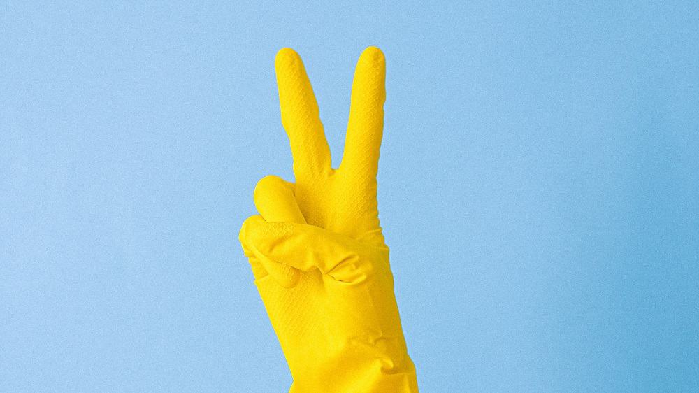 Hand met gele schoonmaakhandschoen doet peace teken op blauwe achtergrond