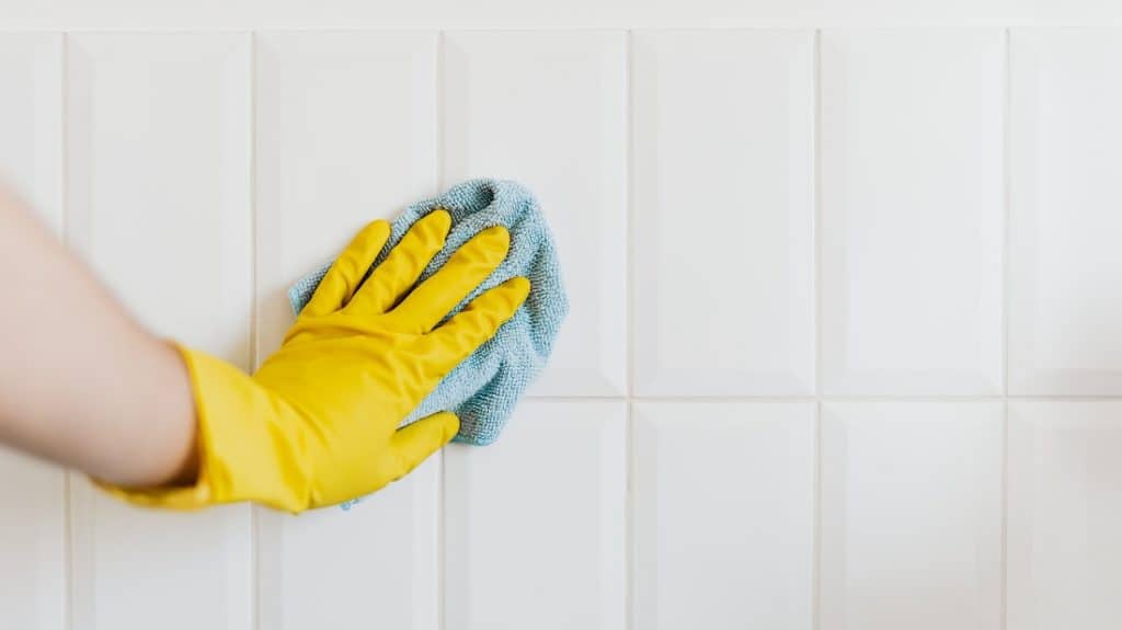 Hand met gele schoonmaakhandschoen maakt tegels schoon met blauwe microvezeldoek