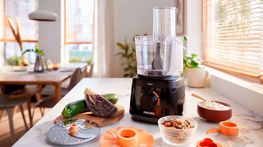een zwarte keukenmachine op tafel met groente en snijbladen