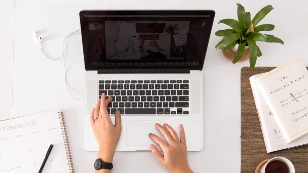 Opengeklapte laptop met grijze behuizing waarop een persoon met een horloge om zijn linkerhand typt. Links van de laptop ligt een agenda. Rechts bovenin staat een plantje.