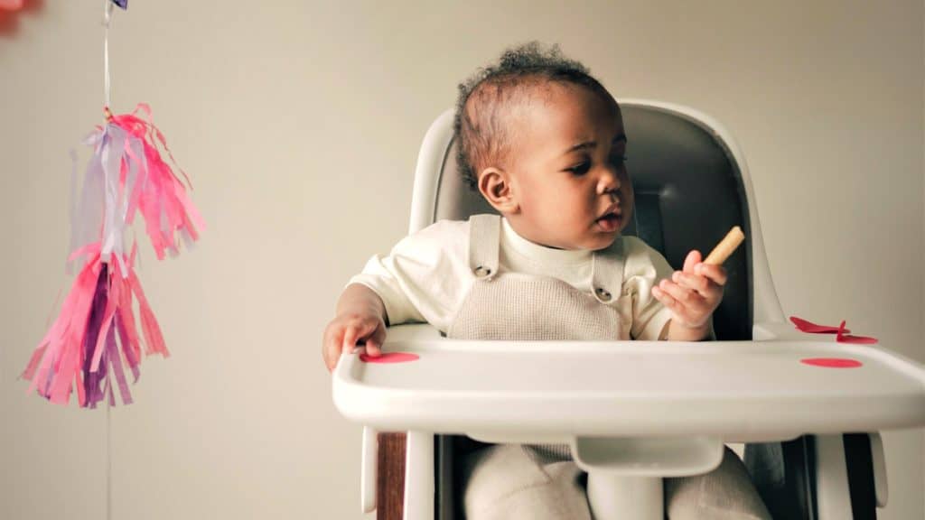 Kindje met een grijze tuinbroek en wit shirtje in een witte kinderstoel. De stoel heeft zwarte bekleding en een eigen eetblad. Links is een paars/roze slinger te zien.