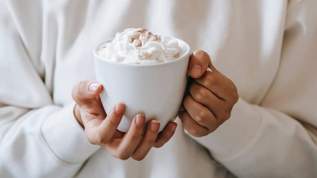 vrouwen handen houden warme drank vast met opgeschuimde melk en cacao of kaneel erover in witte mok