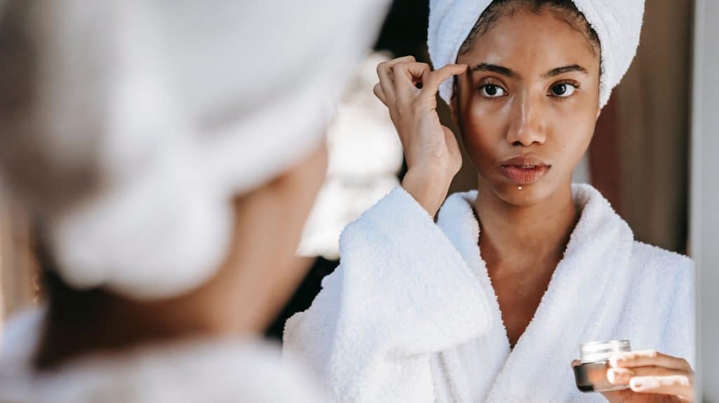 Wazig achterhoofd van vrouw met een witte handdoek over haar haar die in de spiegel kijkt. In de spiegel is de getinte vrouw in een witte badjas te zien met een créme die ze op haar gezicht aanbrengt