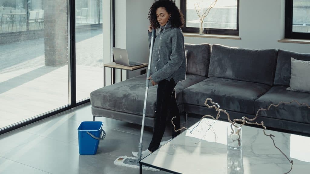 Gekleurde vrouw in comfortabele kleding die een donkergrijze vloer dweilt. Achter haar is een donkergrijze bank te zien met een kussen en voor haar staat een tafel met decoratie. Aan de linkerkant is een glazen wand te zien.