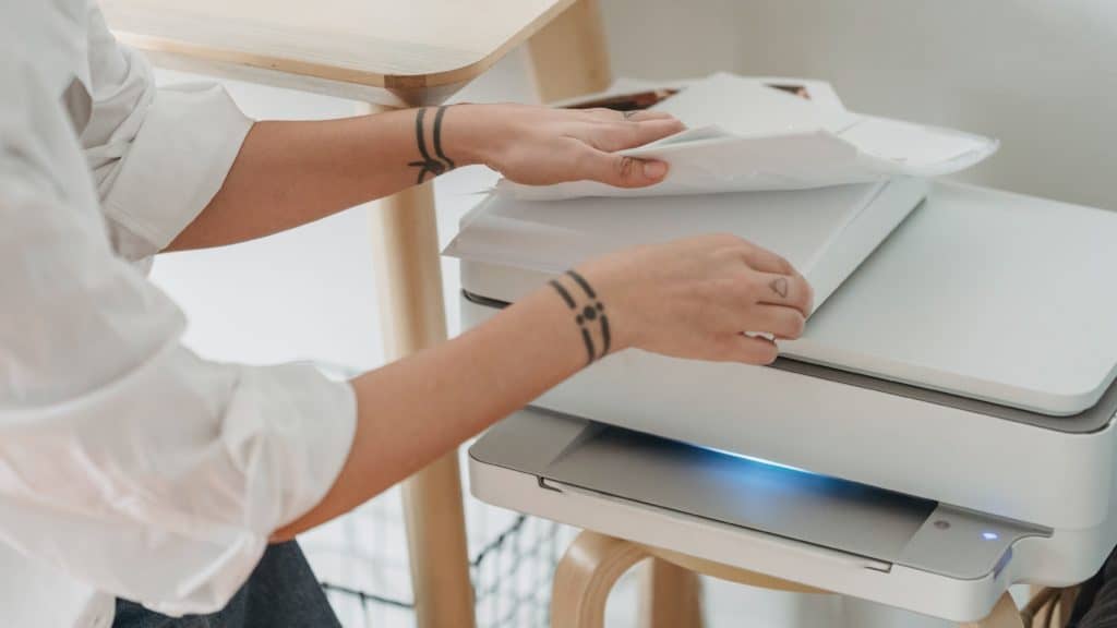 Witte printer op een krukje. Een vrouw die links van de printer staat, bladert door papier wat op de printer ligt.