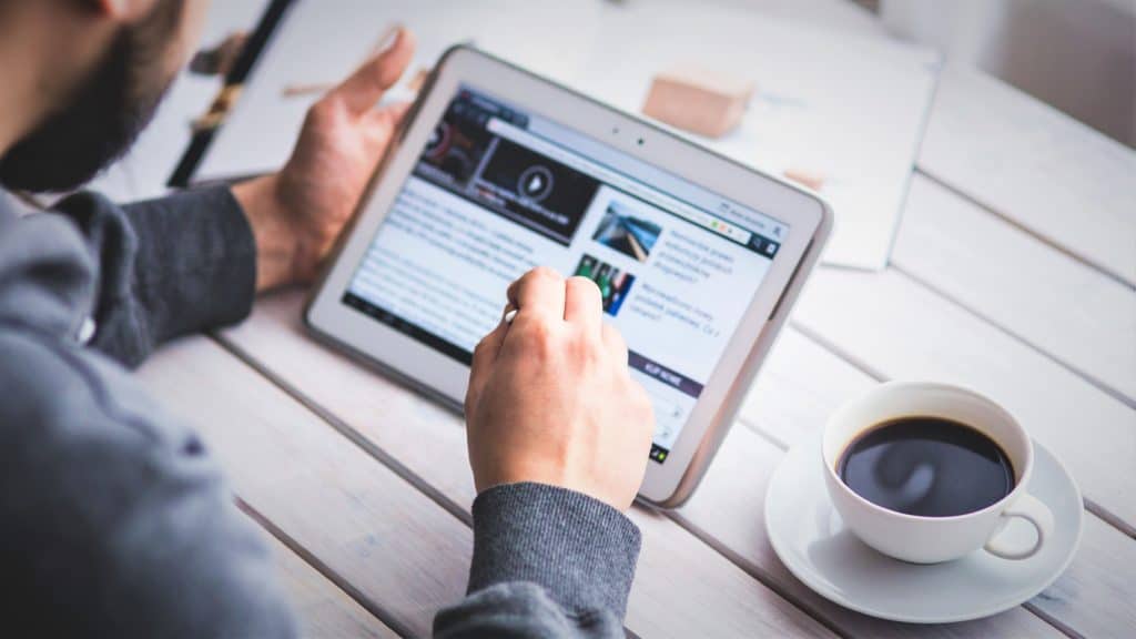 Man die van schuin achter in beeld wordt gebracht terwijl hij met zijn rechterhand met een pennetje op zijnwitte tablet tikt. Zijn linkerhand ondersteunt de tablet Recht van hem staat een kopje koffie.